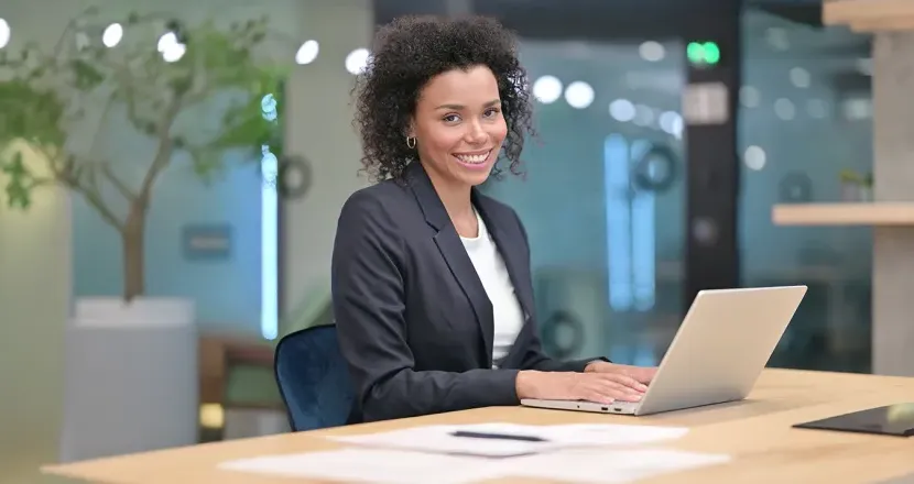 A professional woman sits at a desk with a laptop, smiling confidently, representing career success with a business management degree.